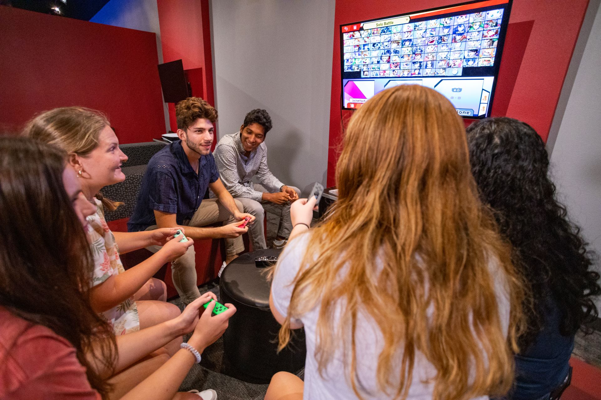 A group of students sitting together in a circle, playing a multiplayer game on a large screen in the Esports center, smiling and interacting with each other while holding gaming controllers